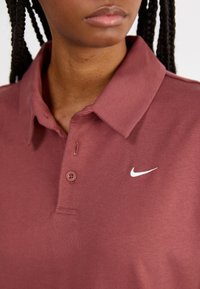 Burgundské polo tričko s límečkem, dvěma knoflíky a bílým logem Nike na levé straně hrudi; hladká textura a bavlněný materiál.