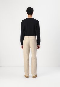 Top negru cu mânecă lungă și pantaloni din in bej, cu buzunare pe spate vizibile și accente crem, asortați cu încălțăminte maro deschis.