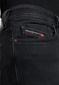 Černé džínové kalhoty s detailním pohledem na zadní kapsu, zdobené červeno-černou štítkovou značkou Diesel Industries a kovovou nýtkou.