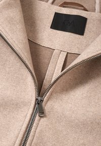 Chaqueta de mezcla de lana beige con cremallera plateada, amplio cuello y una etiqueta negra en el interior. Textura suave con líneas limpias.