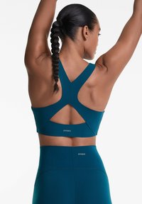 Soutien-gorge de sport teal avec un large design dans le dos, comportant des détails découpés et un ajustement serré. Présenté avec un legging taille haute assorti.