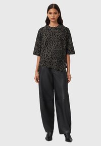 Een vrouw draagt een grijs en zwart luipaardprint t-shirt met korte mouwen, zwarte wijde leren broek en zwarte puntige schoenen, terwijl ze staat.