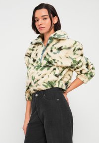 Sudadera de forro polar en color crema con patrones abstractos en verde y marrón, con cuello y cremallera, combinada con pantalones de pana negros.