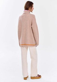 Helles rosa gestricktes Oberteil mit hohem Kragen, orangenem Saumdetail und lockerer Passform, kombiniert mit cremefarbenen Hosen und braunen Schuhen.