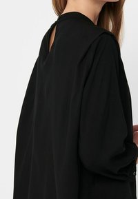 Haut noir à manches longues avec un col montant et une ouverture en forme de clé au dos. Tissu doux, coupe décontractée et poignets avec boutons.