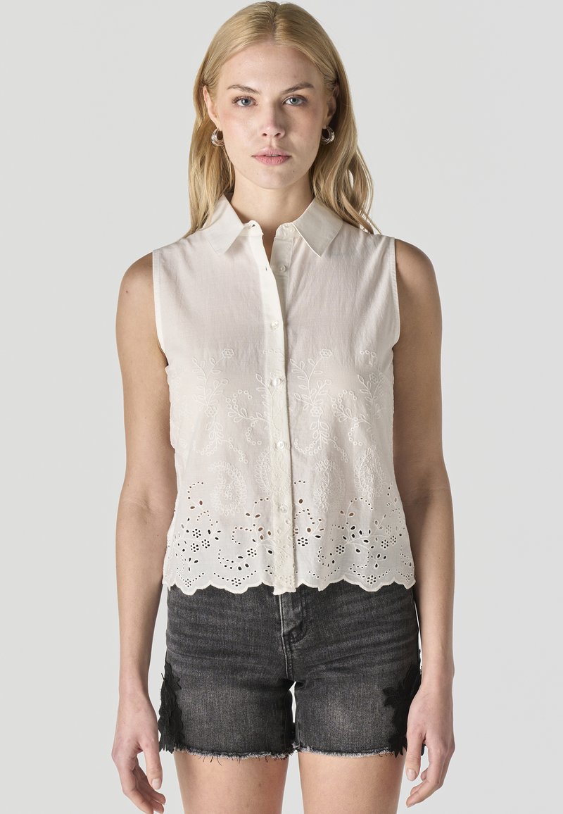 Vrouw draagt een mouwloze witte blouse met knopen en borduursels en zwarte denim shorts met bloemenkantapplicaties, staand tegen een effen achtergrond.