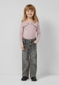 Junges Mädchen steht, trägt ein blassrosa langärmeliges Oberteil mit Schleifendetail, graue Jeans mit weißen Sprenkeln und schwarze Schuhe.