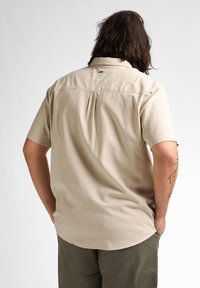Korte mouwen, lichtbeige shirt met een achterste plooi en zachte, gestructureerde stof. Het shirt heeft een klassieke kraag en een subtiele logobezorging bij de hals.