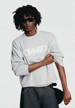 Homme aux cheveux foncés et bouclés avec des lunettes de soleil, portant un sweat-shirt gris clair avec un logo blanc et un jean foncé sur fond uni.