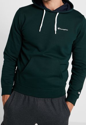 Mann trägt dunkelgrünen Champion-Hoodie mit weißen Kordeln und grauen Jogginghosen, Hände in den Taschen, vor einfarbigem Hintergrund.