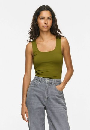 Femme aux cheveux bruns ondulés porte un débardeur vert olive et un jean gris taille haute, une main dans la poche, sur un fond uni.