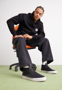 Zapatos negros tipo slip-on con acentos elásticos en los laterales, suelas de goma blancas, combinados con jeans oscuros holgados y una sudadera negra, sentado en una silla naranja.