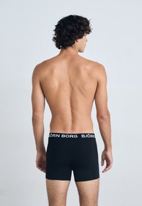 Crni bokserice s bijelim brendiranim pojasom. Tkanina izgleda glatko i prilagođeno, prikazujući elegantan, jednostavan dizajn.