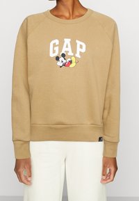 Brązowy sweter z białym napisem "GAP" i grafiką Myszki Miki. Wykonany z miękkiego materiału, z ściągaczami na mankietach i u dołu. Prosty design.