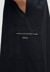 Mână în buzunarul unei haine negre cu un buzunar pătrat cu fermoar, brodat cu cuvântul „obey” în fir alb.