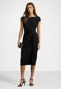 Zwarte jurk met een aansluitende silhoeet, korte mouwen en asymmetrische drapering vastgezet met een decoratieve knoop. Combinatie met zwarte sandalen met bandjes.