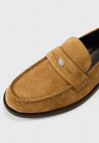 Barna szarvasbőr mokaszín sima textúrával, bőrtalppal és egyszerű pánttal, amelyen ezüst logó díszítés található. Kerek orr kialakítás.