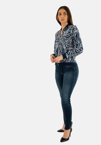 Giacca zip-up blu marina e bianca con un motivo ondulato, abbinata a jeans scuri aderenti e tacchi a punta neri, rivolta di fronte con le mani incrociate.