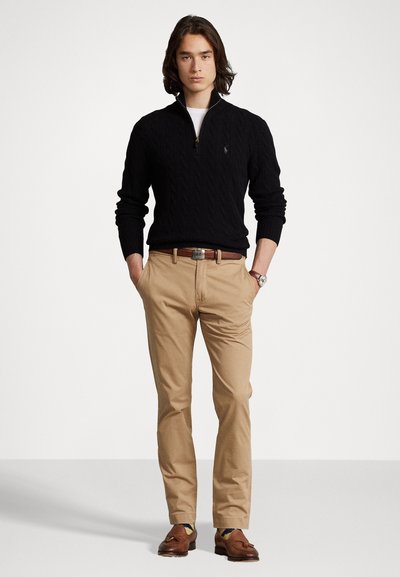 Black cable-knit sweater with a zip collar, beige chinos, brown belt, and loafers. Watch on wrist; model stands with hands in pockets.