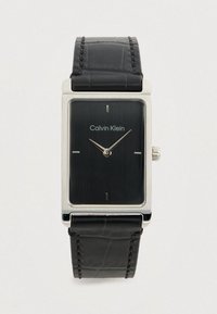 Montre rectangulaire Calvin Klein avec un cadran texturé noir, un boîtier en argent et un bracelet en cuir noir présentant des détails de couture subtils.
