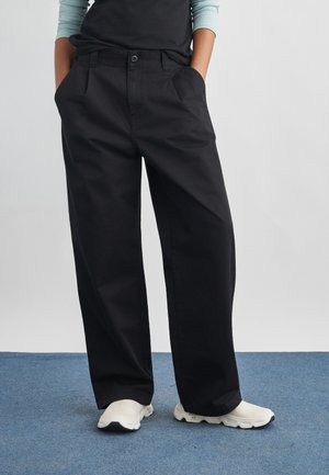 Άτομο που φοράει φαρδιά μαύρα παντελόνια με τα χέρια στις τσέπες και λευκά αθλητικά παπούτσια, στέκεται σε μπλε χαλί μπροστά σε απλό φόντο.