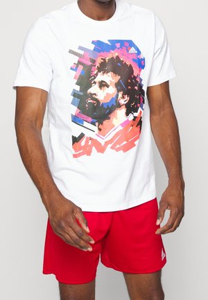 Mężczyzna w białej koszulce Adidas z kolorowym pikselowym portretem mężczyzny i czerwonych spodenek Adidas na jednolitym tle.
