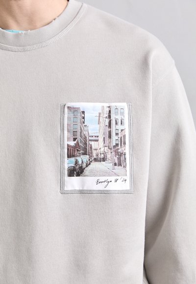 Felpa grigia con tasca rettangolare che presenta un paesaggio urbano stampato e il testo "Brooklyn NY '24." Polsini e scollatura a costine.