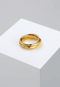 Deux bagues en or lisses et interconnectées reposant sur une plateforme blanche inclinée sur un fond gris.