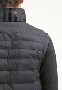 Svart vadderad väst med hög krage, horisontell quiltad design och en unik randig accent på baksidan. Slät textur.