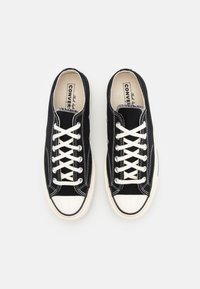 Sorte kanvas sneakers med hvit gummitåhette, med hvite snører og sømmer, og runde lufthull. Flat såle med teksturert grep.