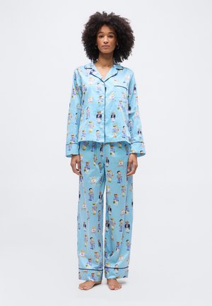 Femeie stând descultă, purtând pijamale cu dungi albastre deschise, cu imprimeuri de personaje de desen animat și cu un top cu mâneci lungi prevăzut cu nasturi.