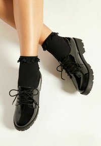 Scarpe stringate in pelle verniciata nera con calze nere texture e bordo arricciato. Il design della suola spessa aggiunge altezza e stabilità.