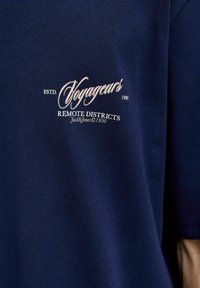 Granatowa bawełniana koszulka z białym haftowanym napisem "Voyageurs ESTD. 1990 REMOTE DISTRICTS" z delikatnym połyskiem.