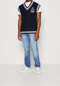 Gilet en tricot à carreaux bleu marine avec col en V, bordure blanche et logo accentué, associé à un jean en denim bleu clair et des baskets blanches.