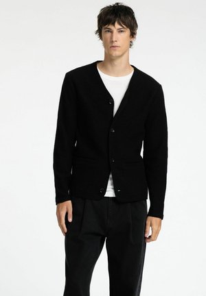 Svart ullblandad cardigan med V-hals, femknappsfront, två framfickor och ribbade manschetter, i kombination med en vit T-shirt och svarta byxor.