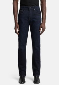 Jeans in denim blu scuro con un taglio dritto, design a cinque tasche e dettagli di cucitura discreti. Indossati con scarpe nere e una maglietta nera.