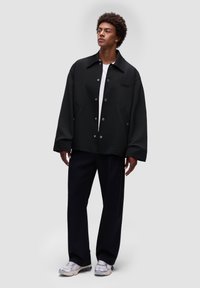 Sort oversized jakke med stor krave, trykknapper og sidelommer, båret over en hvid skjorte, parret med mørke bukser og lyse sneakers.