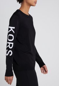 Zwarte sweatshirt met ronde hals, lange mouwen en witte "KORS" tekst op de linker mouw. Gemaakt van zachte, effen stof.