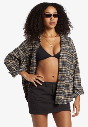 Czarna i żółta koszula w kratę noszona rozpięta na czarnym bikini, zestawiona z czarną mini spódnicą. Posiada długie rękawy i luźny krój.