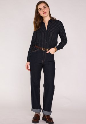 Femme debout, une main dans la poche, portant une chemise noire à fines rayures, un jean foncé retroussé, une ceinture marron et des mocassins marron à glands.