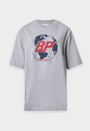 Frau steht mit der Hand in der Tasche, trägt ein graues oversized T-Shirt mit einem Globus und BP-Logo sowie schwarze weit geschnittene Hosen mit kleinen bunten Mustern.