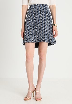 Nainen, joka käyttää sinimusta ruutukuosista hametta, valkoista toppia ja kultaisia korkokenkiä, seisoo yksinkertaisen taustan edessä.