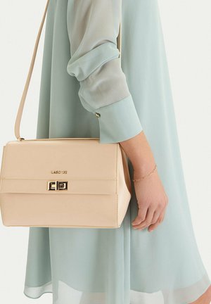 Žena v světle modrých šatech drží béžovou kabelku Lasocki přes rameno se zlatou sponou a náramkem na zápěstí.