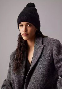 Berretto di lana nera con pom-pom sopra e logo "CK" in rilievo. Indossato con un cappotto grigio testurizzato, caratterizzato da una vestibilità rilassata e design a bavero.