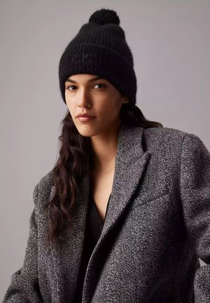 Berretto di lana nera con pom-pom sopra e logo "CK" in rilievo. Indossato con un cappotto grigio testurizzato, caratterizzato da una vestibilità rilassata e design a bavero.