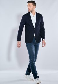 Navy blazer with textured fabric, single button, notch lapels; paired with light blue shirt, dark jeans, and white sneakers.