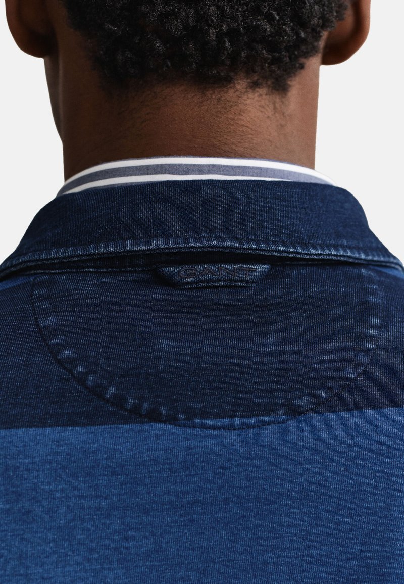 Vista posteriore di una persona che indossa una giacca di jeans scura con un dettaglio cucito a forma circolare sotto il colletto e una camicia a righe sotto.