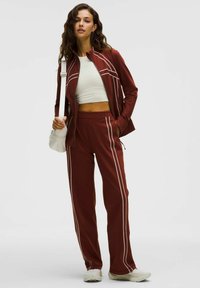 Sarkans sporta uzvalks ar baltām svītrām, kas ietver jaku ar rāvējslēdzēju un bikses ar elastīgu jostasvītu, kombinēts ar baltu krupa krekli un vieglām kurpēm.