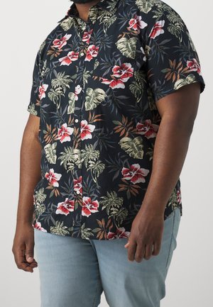 Mann trägt ein kurzärmliges schwarzes Hemd mit roten und weißen Hibiskusblüten sowie grünen tropischen Blättern, kombiniert mit hellblauen Jeans.