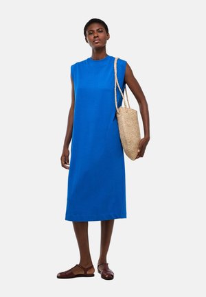 Femme portant une robe midi bleue sans manches, des sandales plates marron et un sac à bandoulière en paille tressée.